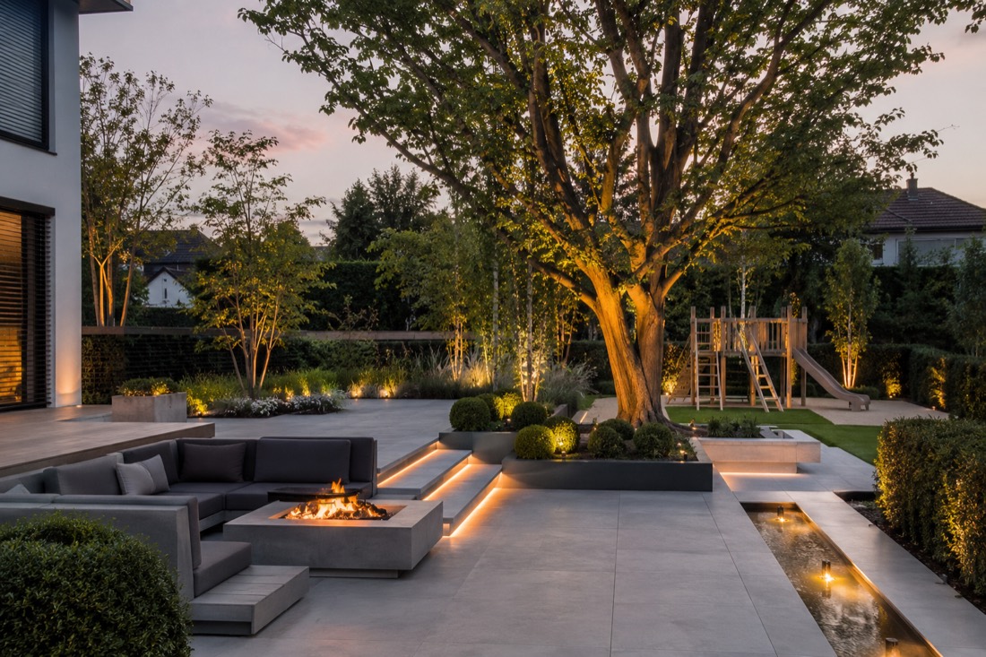 Moderner Garten mit Lounge, Feuerstelle, Wasserlauf und beleuchtetem Baum