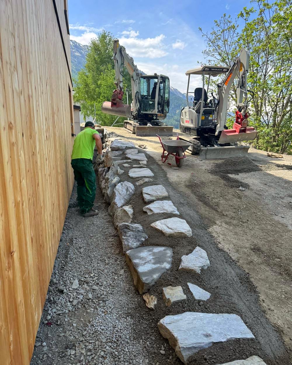 Mitarbeiter setzt eine Natursteinkante entlang einer Holzfassade, dahinter zwei Minibagger